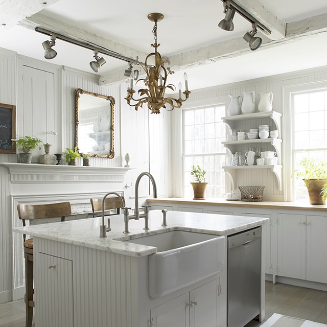 Una acogedora cocina pintada de blanco con paredes con revestimientos de madera y una isla con encimera de mármol y lavabo de estilo granja, estanterías abiertas, una chimenea y ventanas luminosas.