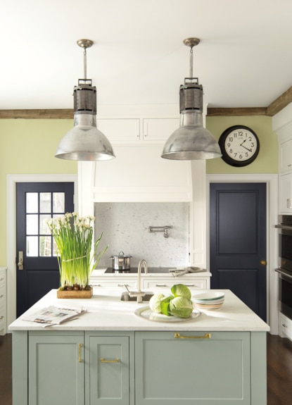 Kitchen with two dark blue doors, white cabinets, green island, modern overhead lighting, and various plants.