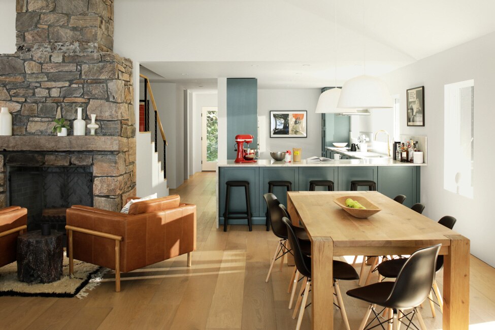 Open-concept living space with midcentury seating near a stone fireplace, wooden dining table, and blue cabinets in a white kitchen. 