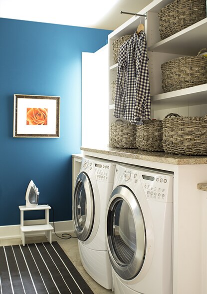 Une salle de lavage présentant un mur bleu, une laveuse et une sécheuse, des étagères blanches, des paniers tressés et un petit marchepied blanc sur lequel est posé un fer à repasser.