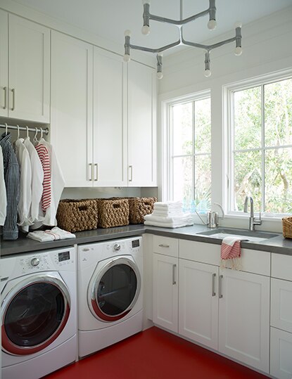 A white-painted laundry room with white washer and dryer, gray countertops, and red floor. Woven baskets, towels and sink on the counter.