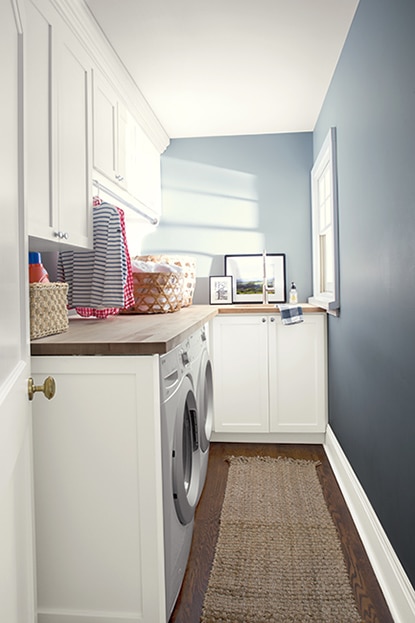 A narrow laundry room with blue walls and light gray-painted trim and ceiling, white cabinetry, washer, dryer, wood countertop and floors and woven baskets.