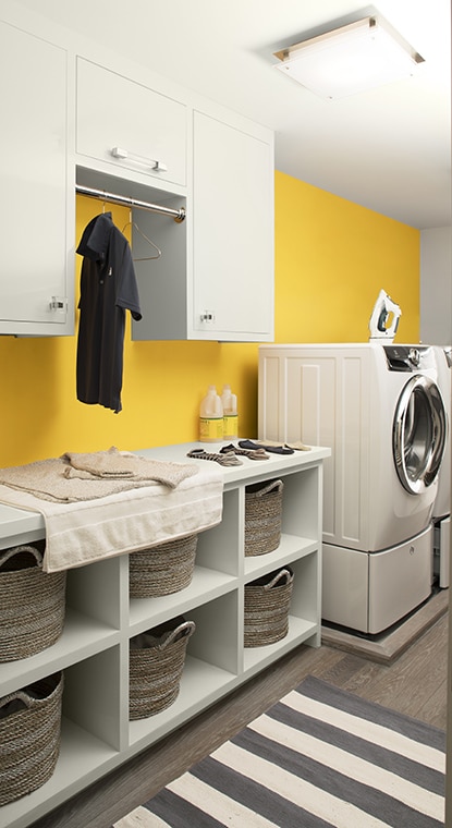 Une salle de lavage présentant un mur jaune, une étagère et des armoires blanches, une laveuse blanche, un plancher en bois gris et un tapis rayé.