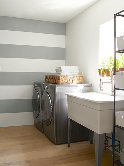 Une salle de lavage blanche comportant un mur d’accent rayé gris et blanc, un évier blanc de style maison de ferme à côté d’une laveuse et d’une sécheuse grises.