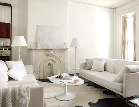 Neutral living room with white walls and an off-white wall mantel, and a white circle coffee table in between two cream couches.