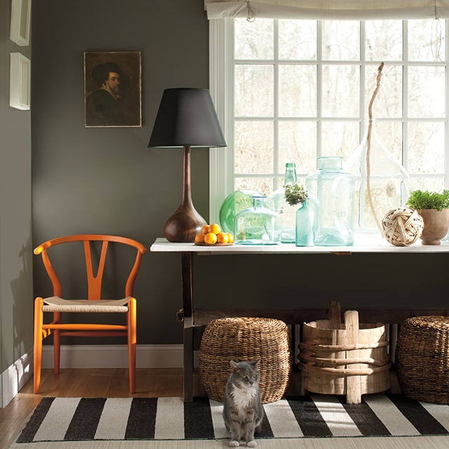 Un rincón de la sala de estar pintado de verde oscuro con marcos blancos, una silla naranja, canastos y una mesa con botellas y una lámpara, frente a un ventanal.