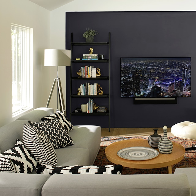 Salon contemporain avec canapé gris, mur d’accent noir avec bibliothèque encastrée, téléviseur grand écran et table basse circulaire.