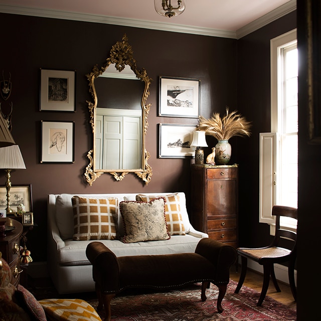 A rich, brown-painted living room with framed artwork and mirror, a loveseat and settee, off-white trim, and a soft pink ceiling.