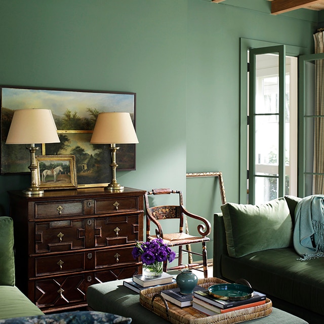 An elegant and relaxing monochromatic living room with rich, green painted walls and open French doors, green velvet sofas, a wood bureau with lamps, and wood beamed ceiling.