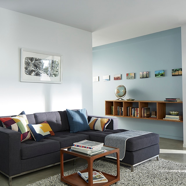 Sun cascading into a light blue and white painted living room with a dark gray sectional sofa and colourful pillows that opens to a light blue hallway wall with a mounted bookcase and framed artwork.