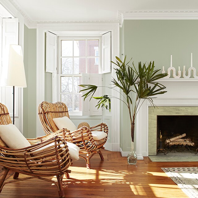 A light green-painted living room wall with beautiful white crown moulding, shutters, trim and fireplace mantel, two rattan chairs with white cushions, and tall green palm leaves in a vase.