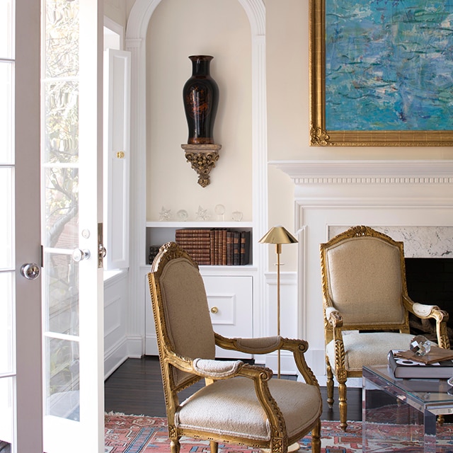 This traditional style living room features neutral tone painted walls with a peach hue, beautiful white decorative moulding, open French doors, turquoise framed art over a white fireplace mantel, and two armchairs.