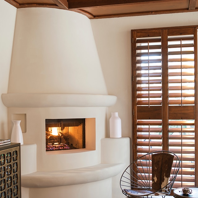 This corner living room space features a beautiful off-white painted adobe style fireplace, off-white walls, a modern metal chair, dark wood window shutters, and a dark wood beamed ceiling.