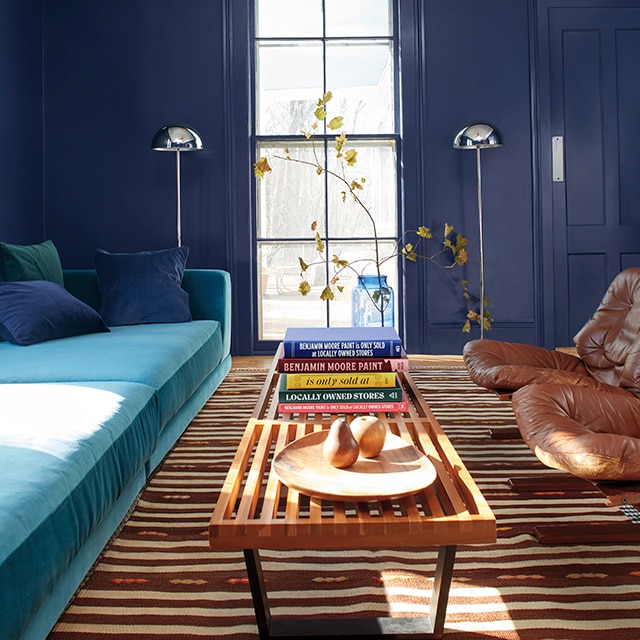 Stunning dark navy blue painted walls and door, and tall centre window are backdrop to this inviting living room space with a turquoise sofa, long wood table, and two leather chairs.