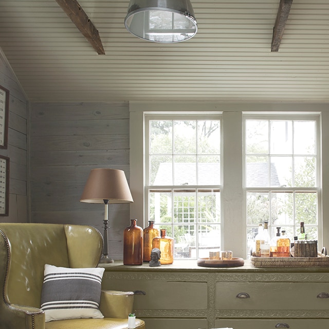A comfortable sitting area with soft, neutral tones, shiplap walls, a light gray-painted beadboard ceiling and trim, a light-sage green dresser under two windows, and a yellow leather chair.