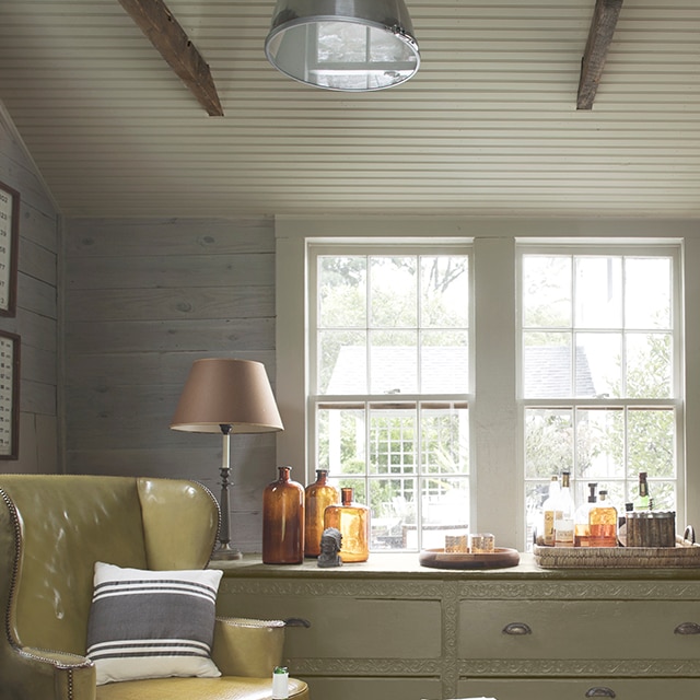 Una cómoda sala de estar con tonos suaves y neutros, paredes traslapadas, marcos y techo de paneles decorativos en gris claro, un tocador verde salvia claro debajo de dos ventanas, y una silla de cuero en color amarillo.