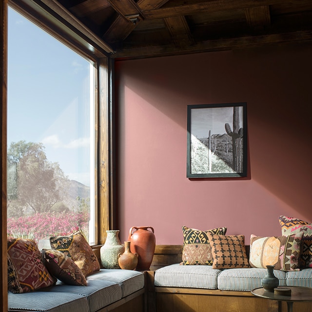 Sun streaming into a muted, brick red-painted living room with a wraparound window seat, southwestern style décor, and a wood beamed ceiling.