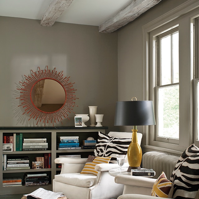 A cozy living room with deep taupe-painted walls, trim, and low built-in bookcase, two white chairs with zebra print pillows, and a white ceiling with wood beams.