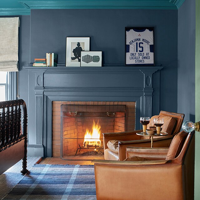 A cozy corner of a room with dark teal-blue painted walls and working fireplace mantel, a teal ceiling and crown moulding, two brown leather chairs, a blue plaid rug, and the edge of a green door.