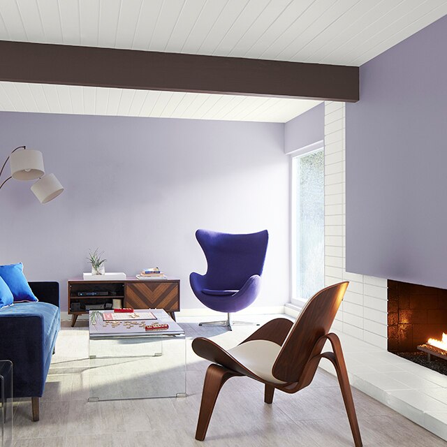 A bright, modern living room with light violet painted walls and accent wall over the fireplace, white shiplap walls and ceiling, a dark brown ceiling beam, navy sofa and purple egg chair.