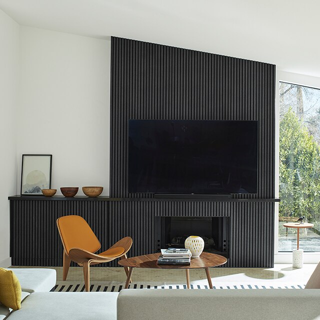 A mid-century modern style living room with bright, white-painted walls and vaulted ceiling, and a black paneled accent wall, built-in cabinet and fireplace, and large screen tv.