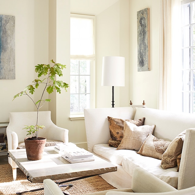 An inviting, sunlit, white-painted living room with a comfy white sofa and two chairs, and a stone top table with a potted plant.