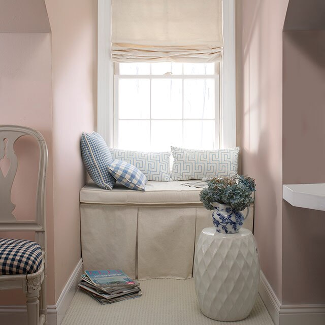 Chambre peinte d’une douce teinte de pêche cendrée avec une fenêtre en alcôve présentant un banc blanc cassé, des coussins décoratifs et une table d’appoint blanche.