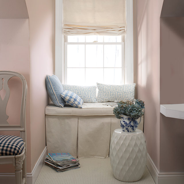 Habitación durazno suave grisáceo con ventana en hueco con un banco color blanco impuro con almohadones y una mesa auxiliar blanca.