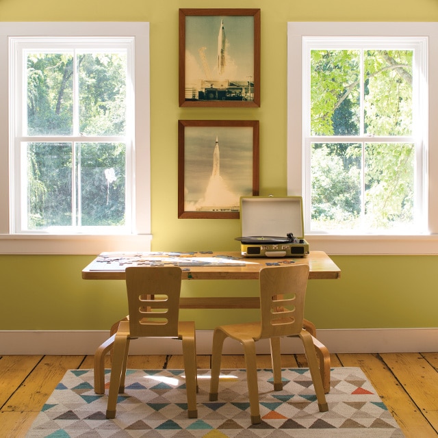 Une table et deux chaises en bois pour enfants avec petit tapis multicolore devant un mur vert pâle avec moulures blanches et images de fusées qu’encadrent deux fenêtres.