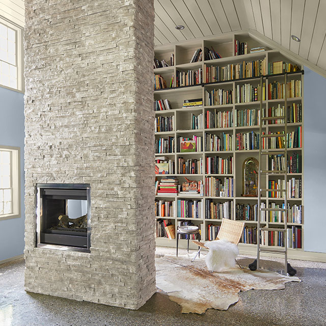 Une salle de lecture à domicile arbore une superbe bibliothèque encastrée pleine longueur de couleur blanche, des murs bleus, un plafond blanc en planches à feuillure, des moulures blanches et une cheminée centrale en pierre blanche.