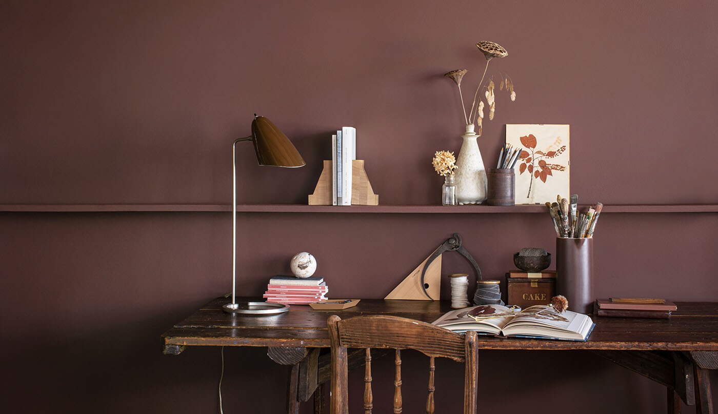 Mur d’un brun intense avec longue étagère suspendue où sont posées différents éléments et bibelots devant une table et une chaise en bois de style rustique.