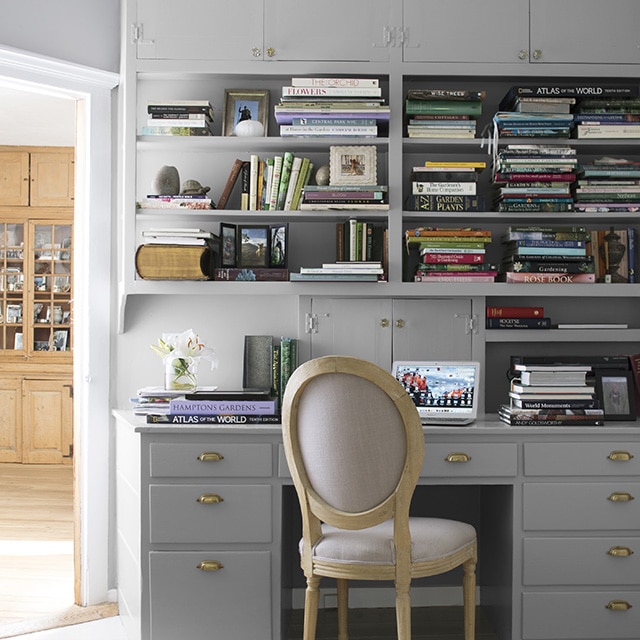 Bureau avec armoires et étagères encastrées grises remplies de livres, table de travail avec plusieurs tiroirs, chaise rembourrée beige et joli luminaire en forme d’étoile.
