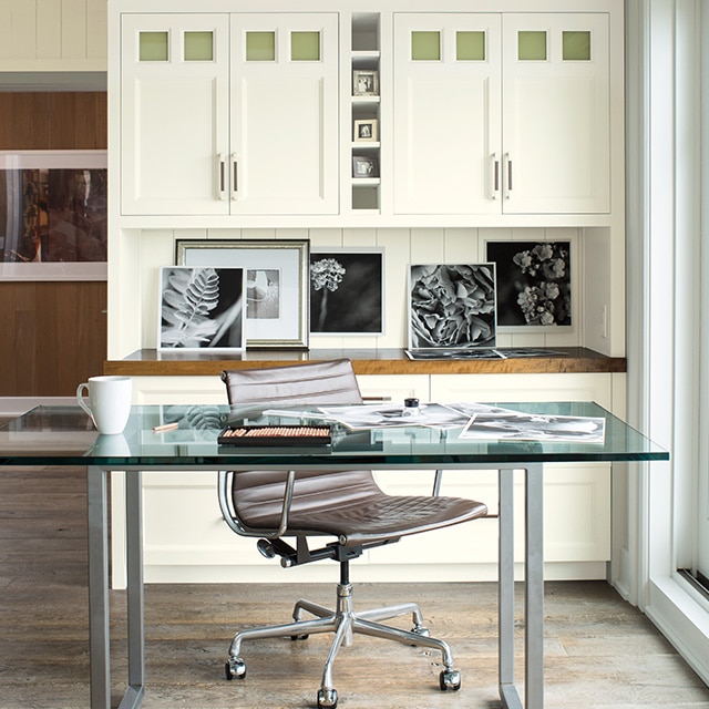 A bright, airy, white painted home office with shiplap walls and white built-in cabinets behind a glass-top desk and brown swivel chair.