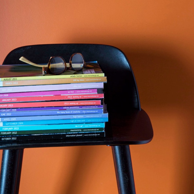 Une chaise noire sur laquelle est posée une pile de magazines de toutes les couleurs se détache du mur orangé.