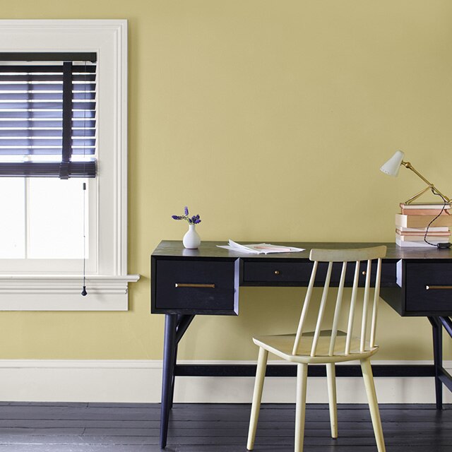 Coin-travail avec bureau noir et chaise jaune, mur jaune pâle, moulures blanches et fenêtre aux stores noirs à moitié relevé.