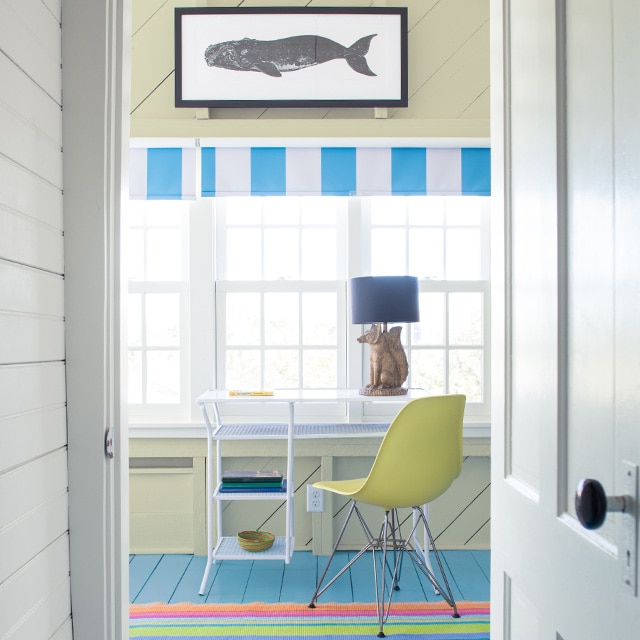 Couloir blanc avec porte blanche qui ouvre sur un bureau à l’ambiance de bord de mer avec mur et plafond en planches à feuillure jaune pâle, tableau de baleine suspendu au-dessus de plusieurs fenêtres, table blanche, chaise jaune et plancher bleu. 