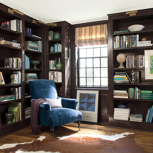 Boudoir brun foncé avec bibliothèques encastrées pleine longueur aux étagères remplies et lumière qui éclaire un fauteuil en velours bleu. 