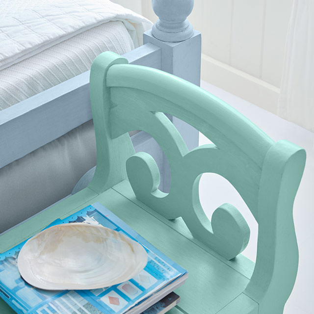The corner of a pale blue-painted wood bed frame and turquoise bench in front of a white wall.