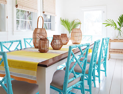 A serene, white-painted dining room with wicker chandeliers, sea blue rattan chairs, a wood table, and yellow striped table runner.