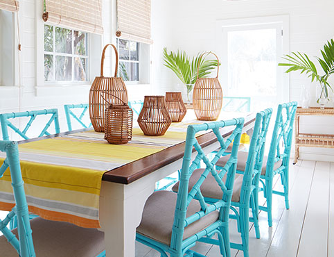 Salle à manger apaisante peinte en blanc avec lustres en osier, chaises bleu océan aux sièges en rotin, table en bois et chemin de table à rayures jaunes.