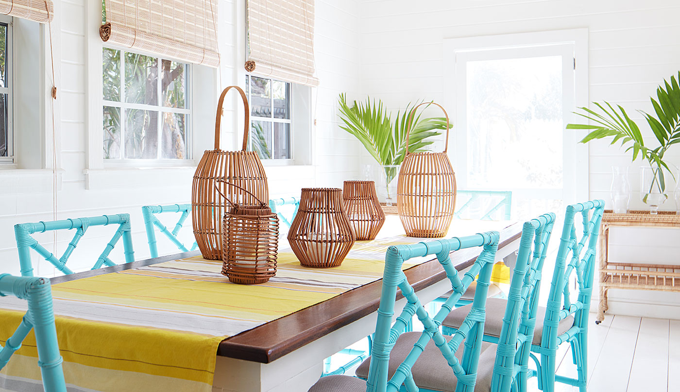 A serene, white-painted dining room with wicker chandeliers, sea blue rattan chairs, a wood table, and yellow striped table runner.