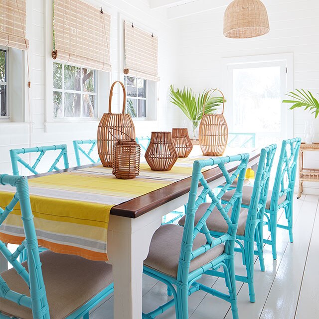 A serene, white-painted dining room with wicker chandeliers, sea blue rattan chairs, a wood table, and yellow striped table runner.