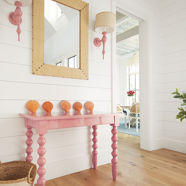 A bright entryway with white-painted shiplap walls, a light pink-painted console, pink-painted wall sconces, and rattan mirror.