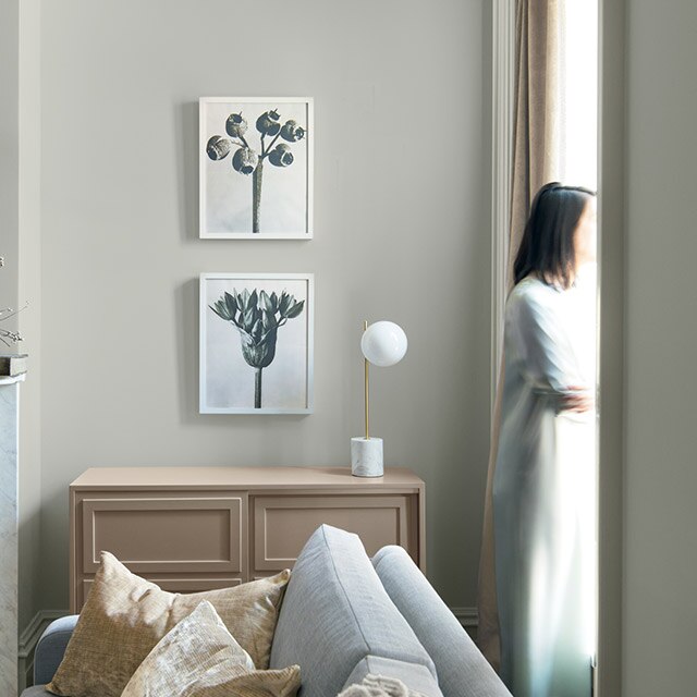 Une pièce accueillante dans des teintes neutres avec des murs gris, une armoire élégante marron clair, un sofa gris et une femme debout près d’une fenêtre.