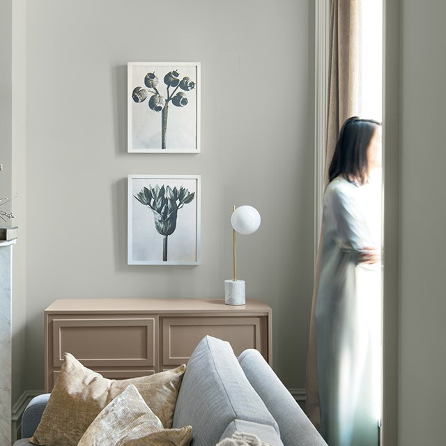 A welcoming room in neutral hues and gray-painted walls, an elegant tan cabinet, gray sofa, and a woman standing near a window.