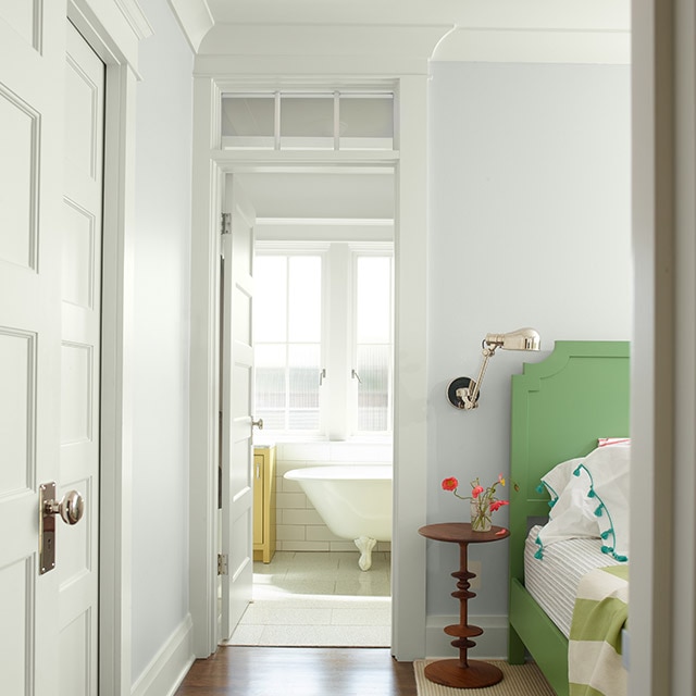 A welcoming white-painted bedroom with green headboard, green and white bedding, a small wooden side table, and a door open to claw-foot tub in a white bathroom.