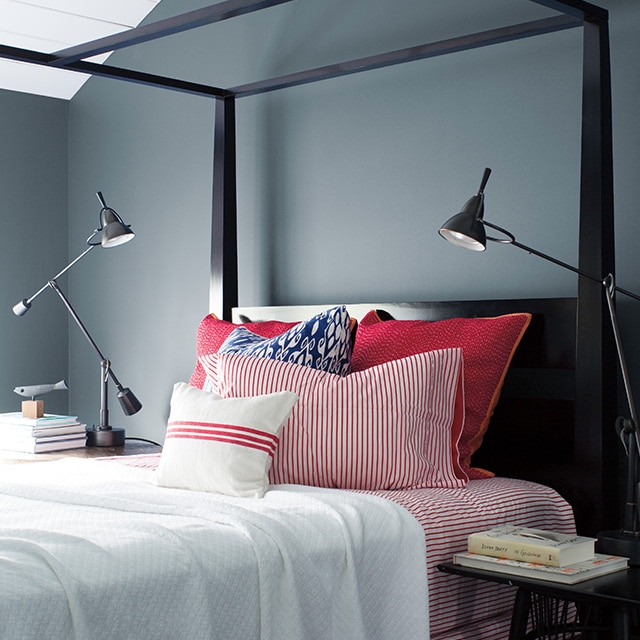 Sleek, Wolf Gray-painted bedroom with black bedframe, red and white bedding, two swing-arm lamps, and stacks of books.