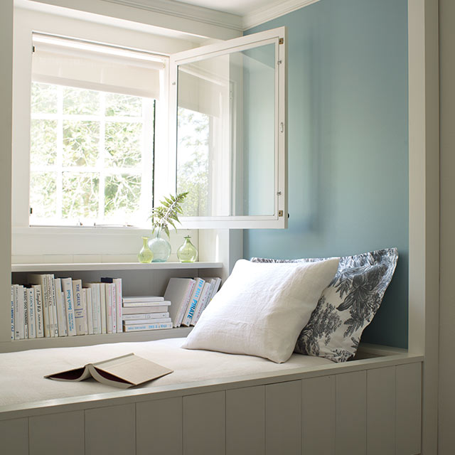 Une chambre blanche présentant un mur d’accent peint en bleu derrière un coin lecture intégré et une fenêtre ouverte.