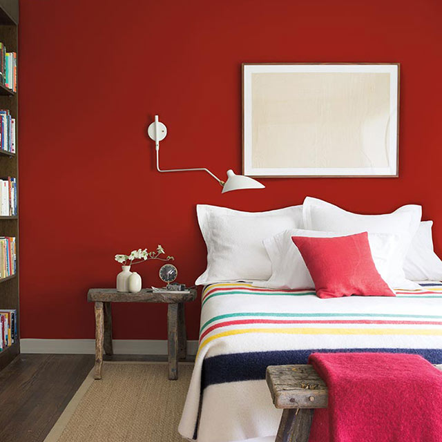 Bedroom with red-painted walls, white trim, black bookcase, colourful bedding, and wooden accent tables.