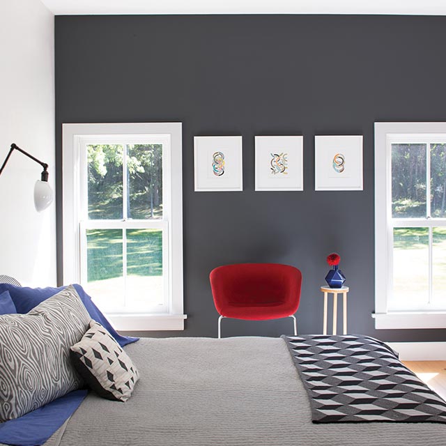 A bedroom with a dark gray accent wall, red chair, gray bedding, and wall art.